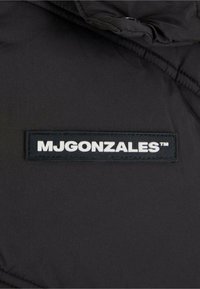 Schwarze gepolsterte Jacke mit glatter Textur, ausgestattet mit einem rechteckigen, erhabenen schwarzen Etikett mit weißem Text: "MJGONZALES™."