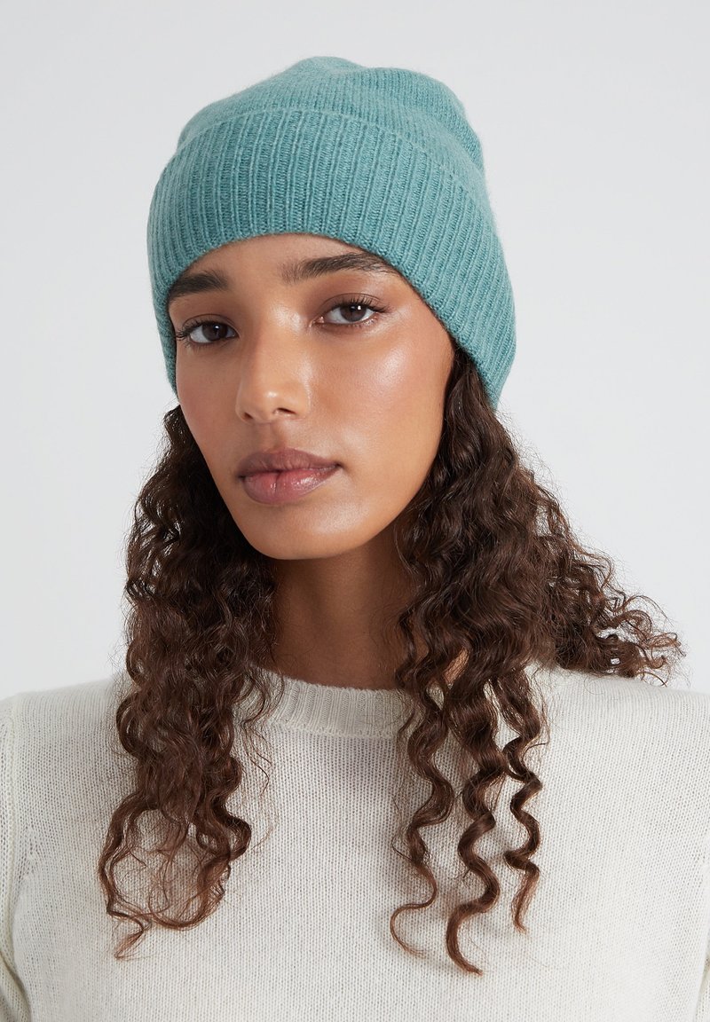 Un berretto a maglia coste color verde acqua, texture morbida, vestibilità aderente, indossato con un maglione di colore chiaro. Design minimalista con una silhouette pulita.