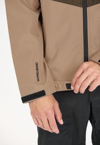 Brun vandtæt jakke med en mørkegrøn panel, justerbare velcro-vrængninger og en sort lynlås, med teksten "WPRO 8000" på ærmet.