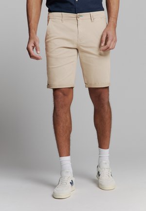 Beige shorts with rolled hems, made of cotton blend fabric. Featuring a button closure and side pockets. Worn with white socks and sneakers.