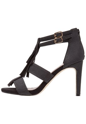 Sort højhælet sandal med teksturerede stropper, dobbelte spænder, frynse detaljer og lynlås lukning bagpå.