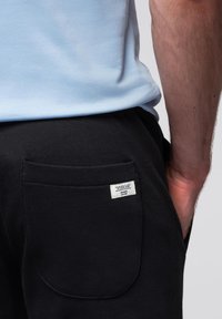 Zwarte broek met een achterzak met een wit label. De stof heeft een gladde textuur. Een lichtblauw shirt is zichtbaar boven de tailleband.