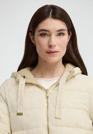 Junge Frau mit langen braunen Haaren trägt eine cremefarbene, gesteppte Jacke mit Kapuze und goldenen Creolen, neutraler Hintergrund.