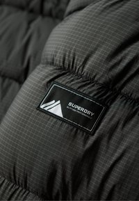 Detalle de una chaqueta de plumas negra que presenta una etiqueta cosida. La textura del material es acolchada con un patrón de cuadrícula; el logotipo incluye gráficos de montañas y texto.