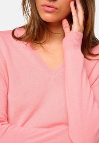 Femme portant un pull en V rose pâle, touchant le côté de son visage avec la main, montrant une partie de ses lèvres et ses cheveux bruns.