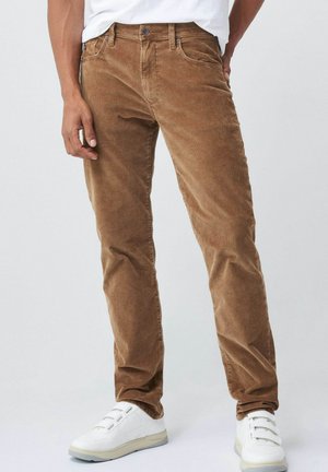 Man wearing brown corduroy pants and white sneakers standing against a plain background.