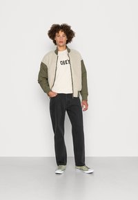 Bomber jacket with a cream fleece body and olive green sleeves, paired with a light beige T-shirt featuring bold black text, black jeans, and green sneakers.