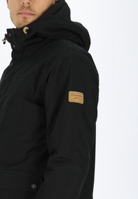 Black waterproof jacket with a streamlined design, featuring a hood, snap buttons, and a tan label on the left sleeve.