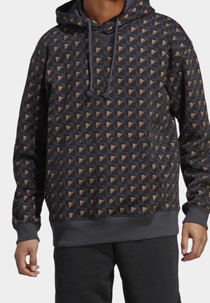 Mann trägt schwarzen Kapuzenpullover mit Muster aus kleinen goldenen Adidas-Logo-Dreiecken und gerippten Bündchen und Saum.