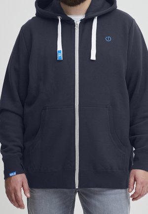 Personne portant un sweat à capuche bleu marine zippé avec des cordons blancs et un jean gris clair, debout devant un fond uni.