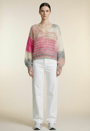Femme debout face à l’avant portant un pull à rayures duveteux col en V rose et gris, un pantalon blanc et des baskets blanches sur un fond uni.
