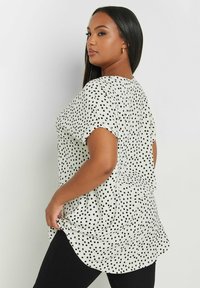 Blusa a pois bianca e nera con maniche corte, tessuto morbido, vestibilità fluida e orlo leggermente curvo. Motivo testurizzato su tutta la superficie.