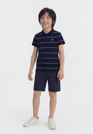 Usmievajúci sa chlapec stojaci, oblečený v námornícky modrom pruhovanom polo tričku, námorníckych šortkách a bielych teniskách na jednoduchom bielom pozadí.