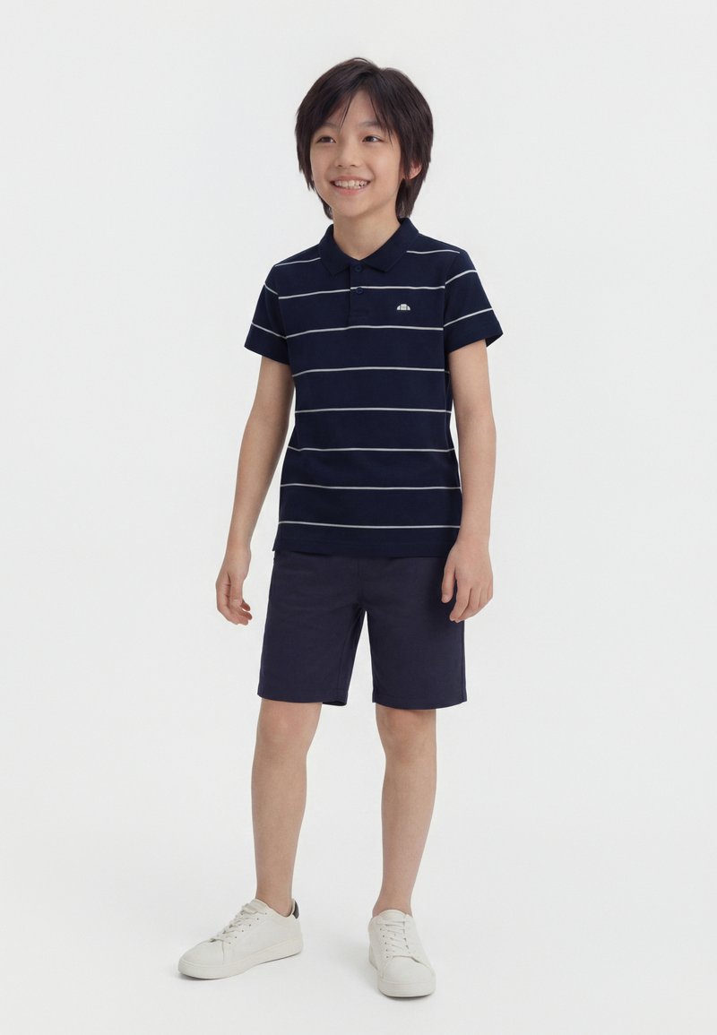 Garçon souriant debout portant un polo rayé bleu marine, un short bleu marine, et des baskets blanches sur un fond blanc uni.