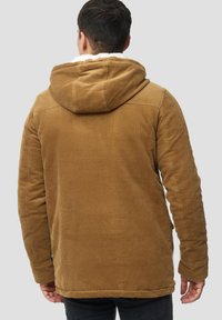 Hombre con una chaqueta de pana marrón con capucha y forro blanco, visto desde atrás sobre un fondo gris liso.