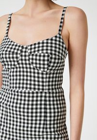 Schwarzes und weißes Gingham-Kleid mit dünnen Trägern, einem taillierten Oberteil mit zwei Taschen und einer glatten Stoffoberfläche.