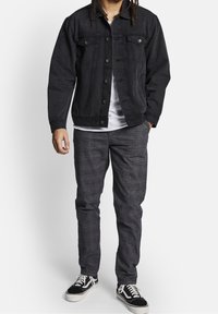 Zwarte denim jas met knoopsluiting, twee borstzakken, gecombineerd met grijs geruite broek en zwarte canvas sneakers.