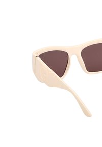 Beige zonnebril met een geometrische vorm, donkerbruine lenzen en een reliëflogo op de poot. Het materiaal lijkt glad en glanzend.
