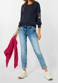 Felpa navy con ricami floreali sulle maniche, abbinata a jeans azzurri arrotolati e scarpe da ginnastica bianche, con una sciarpa rosa in mano.