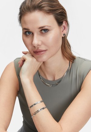 Schmuck in Silberton präsentiert eine elegante, strukturierte Halskette mit zwei Schichten und ein passendes Armband an einem Arm, vor einem neutralen Hintergrund.