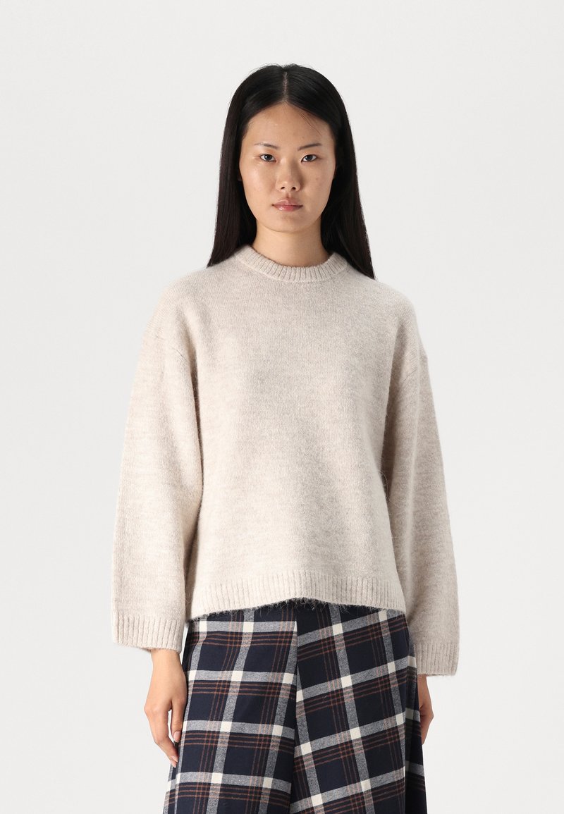 Ανοιχτό μπεζ πλεκτό πουλόβερ με στρογγυλή λαιμόκοψη και άνετη εφαρμογή. Διαθέτει ριγέ μανσέτες και στρίφωμα, συνδυασμένο με φούστα καρό.