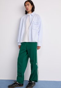 Lysblå stribet skjorte med knaplukning, lagd ovenpå en hvid T-shirt, parret med grønne træningsbukser med hvide sidelæg og sorte sneakers.