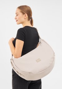 Borsa a spalla beige realizzata in tessuto morbido, con una forma arrotondata, tracolla regolabile e una piccola etichetta con logo sul davanti.