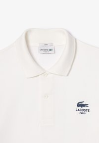Bílá polo košile vyrobená z texturované látky, s klasickým límcem, tříknoflíkovým zapínáním a námořnickým logem Lacoste na levém prsu.