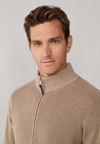 Beige geribbelde trui met een hoge kraag en ritssluiting, voorzien van een gestructureerde stof en subtiele stikdetails op de schouders.