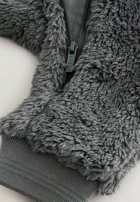 Sivá chlpatá bunda s textúrovaným, chlpatým vonkajškom, hladkým vnútrom a elastickými manžetami. Detail so zipsom je výrazne viditeľný.