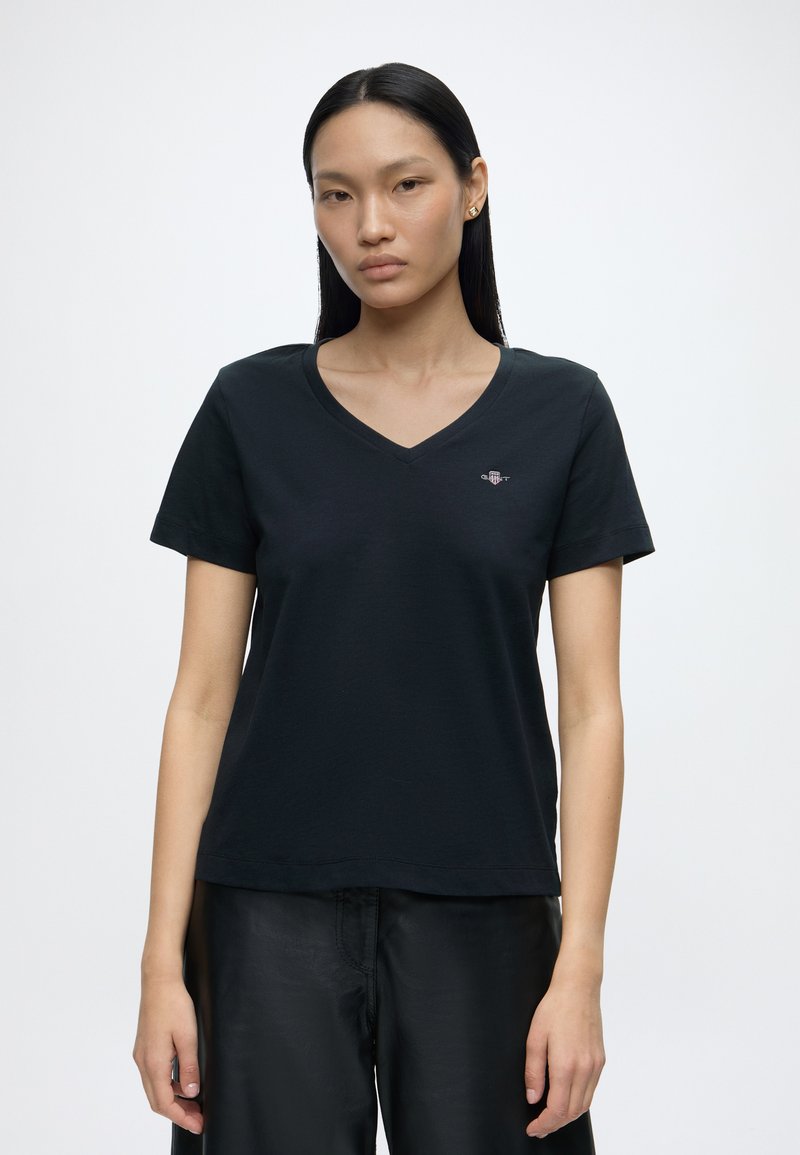 Femme aux longs cheveux foncés portant un T-shirt noir col en V avec un petit logo sur la poitrine et un pantalon noir, debout devant un fond blanc uni.