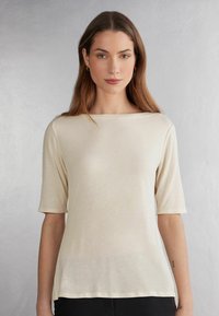Lyse beige kortærmet top med bådudskæring, lavet af blødt stof. Har en afslappet pasform og glat overflade, parret med sorte bukser.