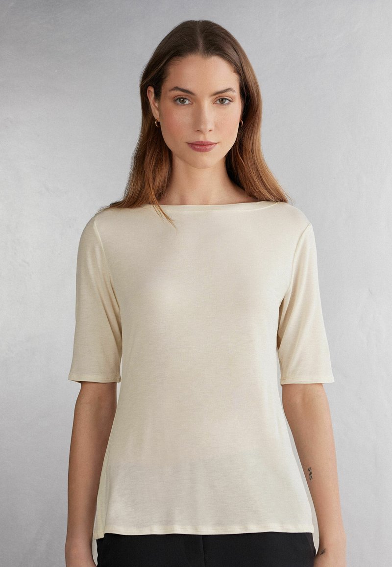 Lyse beige kortærmet top med bådudskæring, lavet af blødt stof. Har en afslappet pasform og glat overflade, parret med sorte bukser.