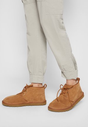 Personne portant un pantalon beige clair et des bottines en daim marron clair à lacets avec semelles en caoutchouc, montrée de la moitié du mollet vers le bas sur un fond uni.