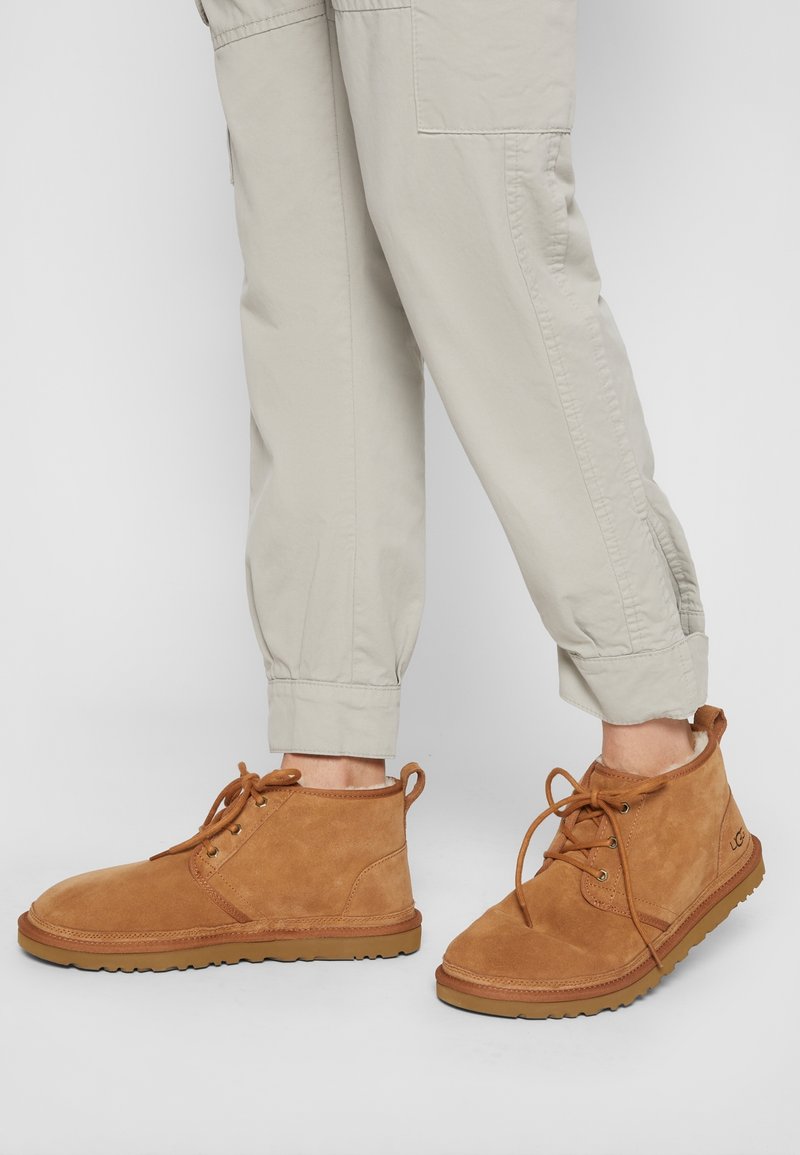 Personne portant un pantalon beige clair et des bottines en daim marron clair à lacets avec semelles en caoutchouc, montrée de la moitié du mollet vers le bas sur un fond uni.