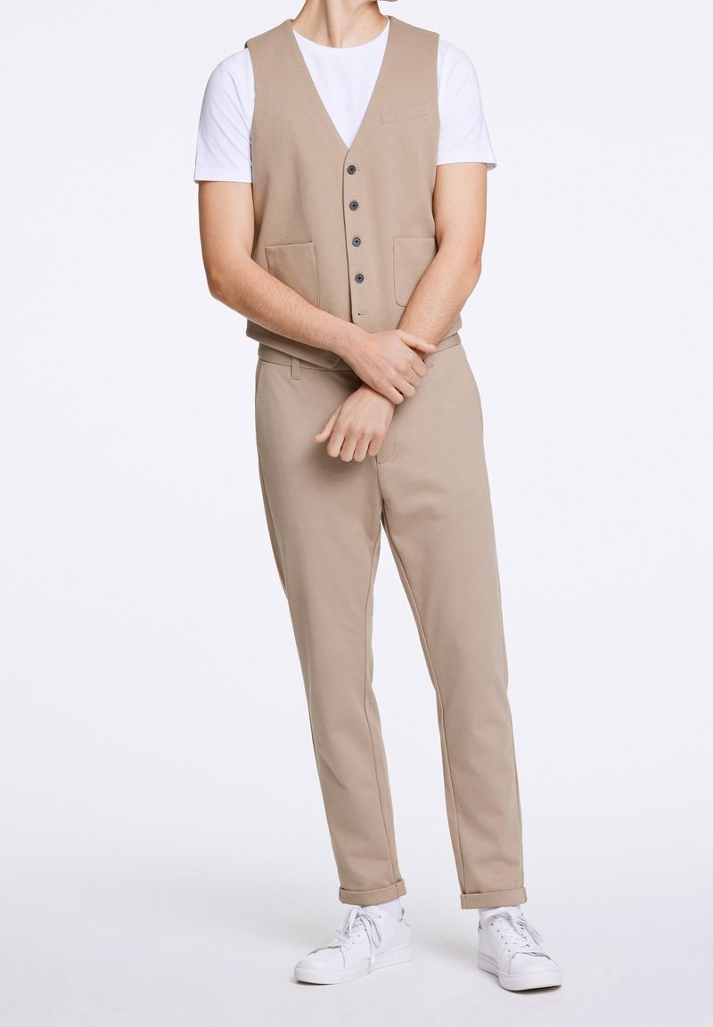 Homme portant un gilet beige boutonné, un pantalon assorti aux ourlets retroussés, une chemise blanche à manches courtes, et des baskets blanches, debout les bras croisés.