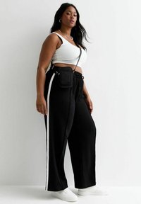 Zwarte wijde broek met witte zijstrepen, gecombineerd met een witte crop top. Accessoires omvatten een kleine zwarte schoudertas en witte sneakers.