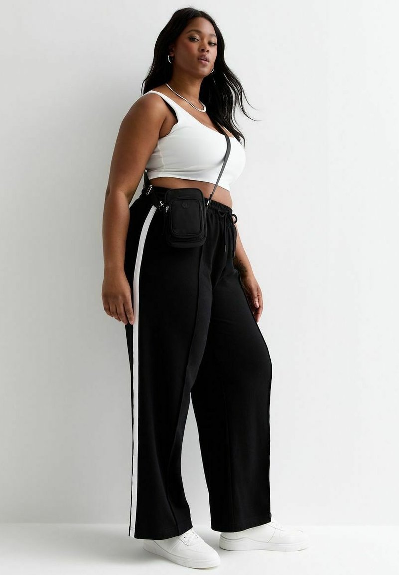 Zwarte wijde broek met witte zijstrepen, gecombineerd met een witte crop top. Accessoires omvatten een kleine zwarte schoudertas en witte sneakers.