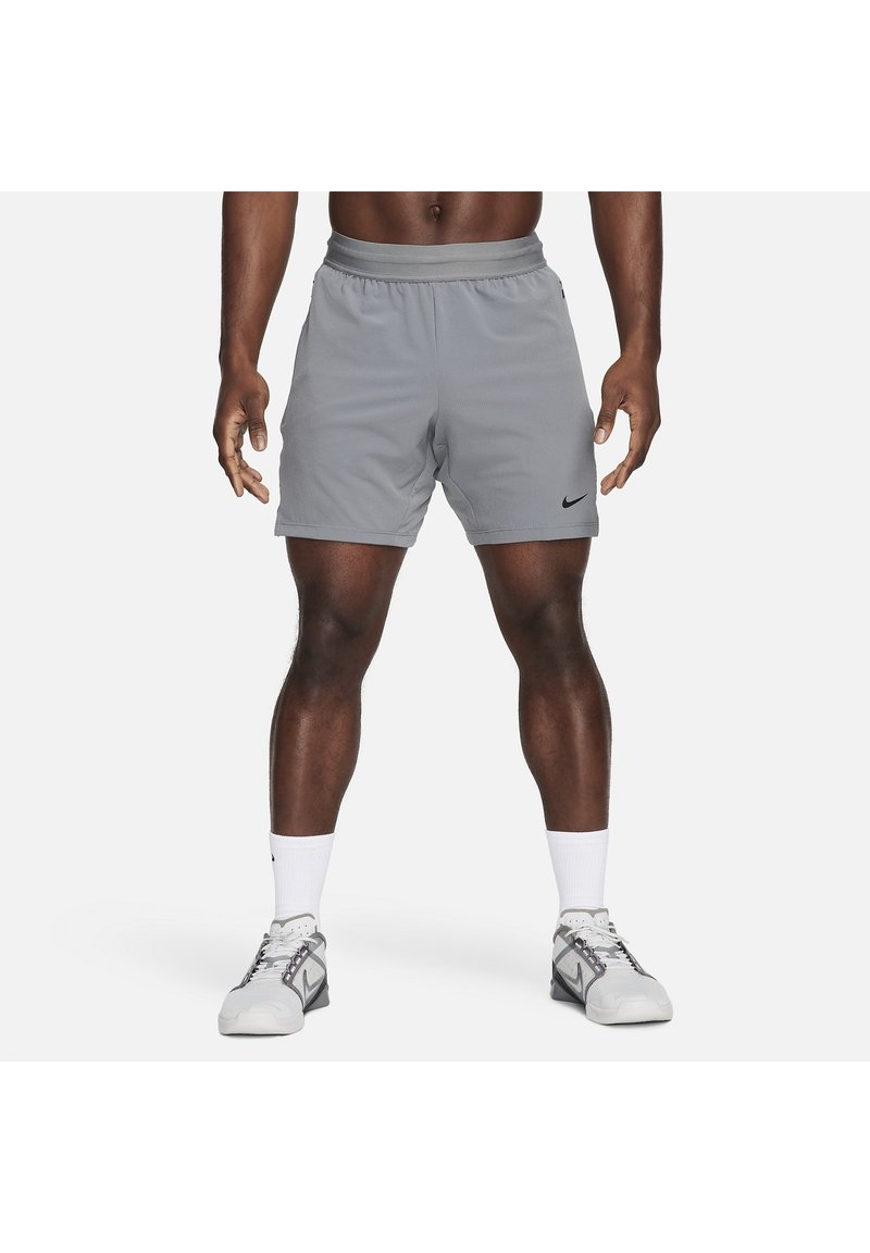 Homme portant un short de sport gris avec un logo Nike noir, des chaussettes blanches et des baskets Nike grises, debout sur fond blanc.