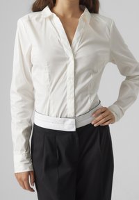 Chemise blanche à manches longues avec col et fermeture à boutons ; associée à un pantalon noir taille haute avec une ceinture blanche à coutures contrastées.
