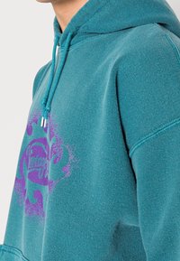 Tyrkysová mikina s kapucí, zdobená fialovým vintage grafickým motivem. Vyrobena z texturované látky s nastavitelnými šňůrkami.