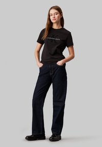 Camiseta de algodón negra con el logo "Calvin Klein Jeans" en verde, combinada con jeans de denim de pierna ancha azul oscuro y botas negras hasta el tobillo.