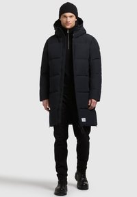Czarna puchowa kurtka z kapturem, z zapięciem na zamek, pikowanym wzorem i bocznymi kieszeniami. Noszona na czarnych spodniach i butach.