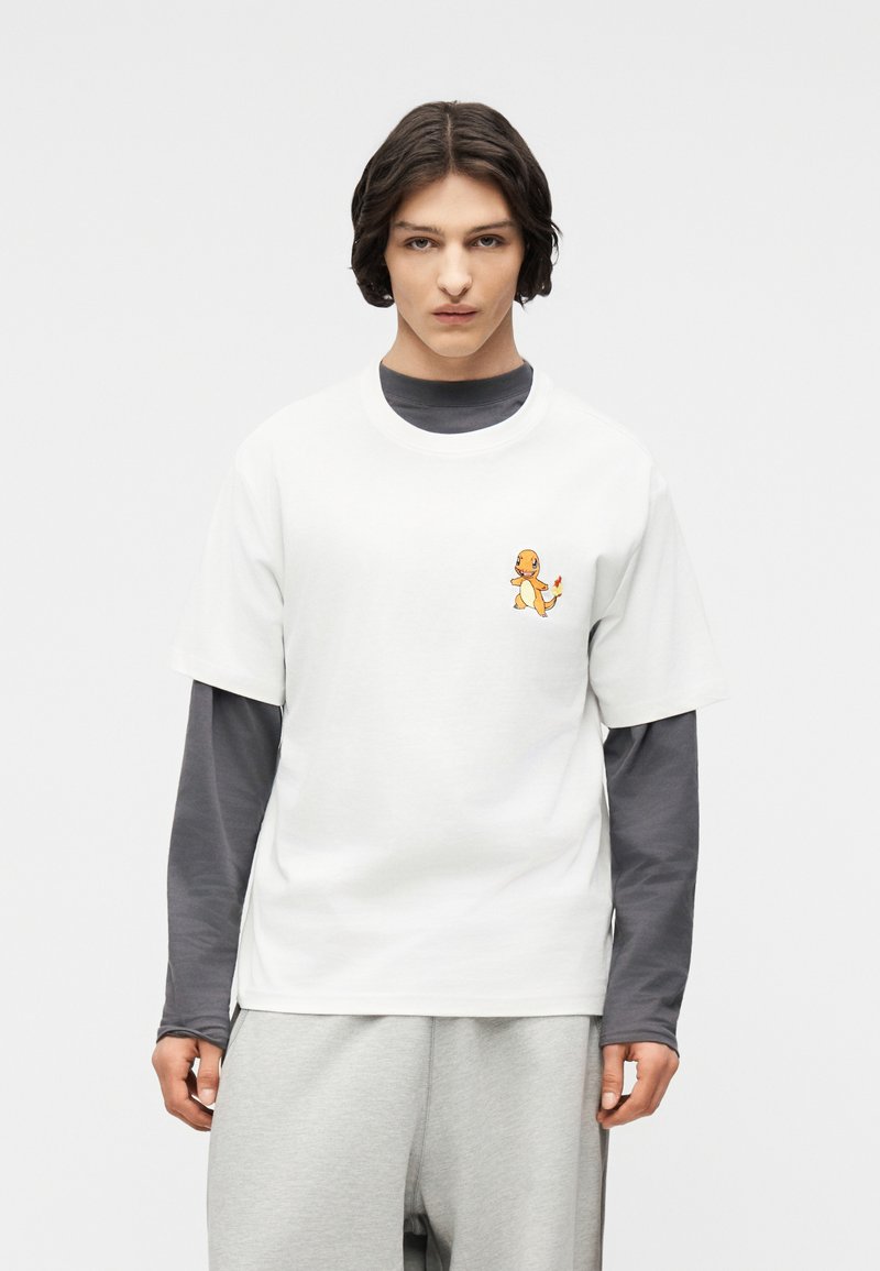 Νεαρό άτομο με σκουρόχρωμα μαλλιά που φοράει ένα λευκό T-shirt με ένα μικρό γραφικό Charmander πάνω από μια γκρι μπλούζα μακρυμάνικη και ανοιχτό γκρι παντελόνι.