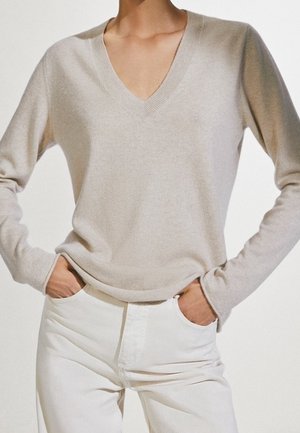 Person wearing light beige V-neck long-sleeve sweater with hands in pockets of white pants against a plain background.