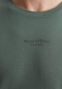 Groene katoenen T-shirt met een ronde hals; voorzien van marineblauwe bedrukte tekst "Marc O'Polo EST. IN STOCKHOLM" op de voorkant.