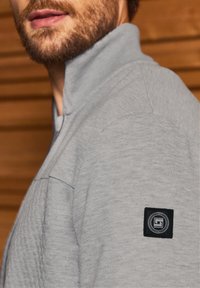 Pull gris avec col montant, présentant un motif texturé sur l'épaule et un patch logo carré noir sur la manche.