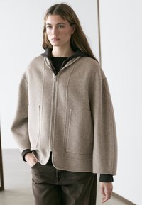 Junge Frau mit langen Haaren, die eine beige Jacke mit Reißverschluss über einem dunklen Hoodie und dunkler Hose trägt und vor einem schlichten Hintergrund drinnen steht.