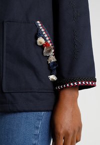 Marinblå jackärm med broderad kursiv text, dekorativa fransar och röd, vit och svart flätad kant ovanför en mörkhyad hand.