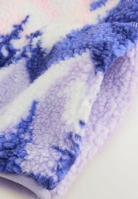 Pătură din fleece cu o suprafață moale și texturată în nuanțe de violet, alb și roz deschis, având un model asemănător cu nori. Marginea este finisată cu atenție.